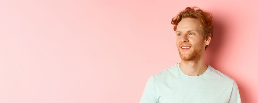 Portrait of attractive caucasian guy with red messy hair and beard, turn head and looking left with pleased smile, standing over pink background.