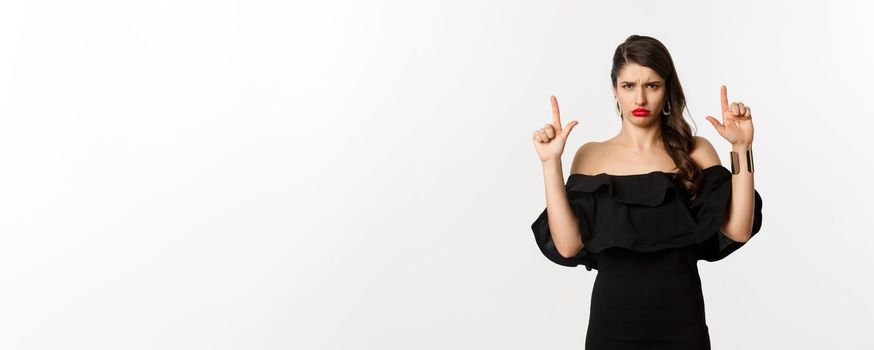 Fashion and beauty. Disappointed woman sulking upset, pointing fingers up and complaining, standing dissatisfied in black dress, white background.