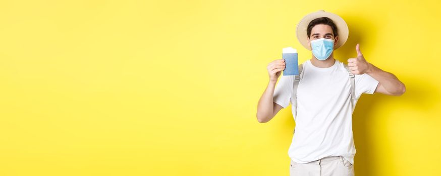 Concept of covid-19, tourism and pandemic. Happy male tourist in medical mask showing passport, going on vacation during coronavirus, make thumb up sign, yellow background.