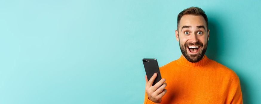 Close-up of happy handsome man writing message on mobile phone, holding smartphone and smiling, standing against turquoise background.