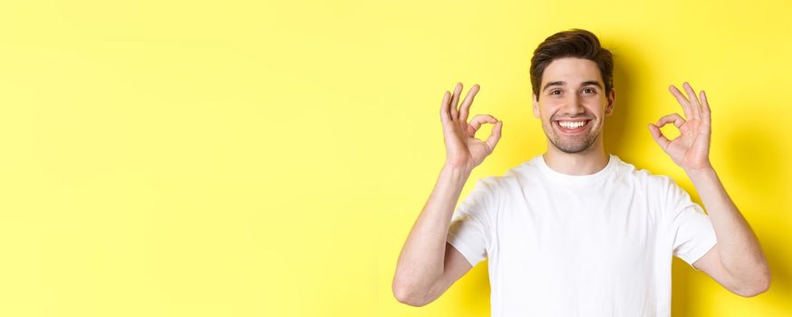 Close-up of handsome young man showing okay sign, approve and agree, smiling satisfied, standing over yellow background.