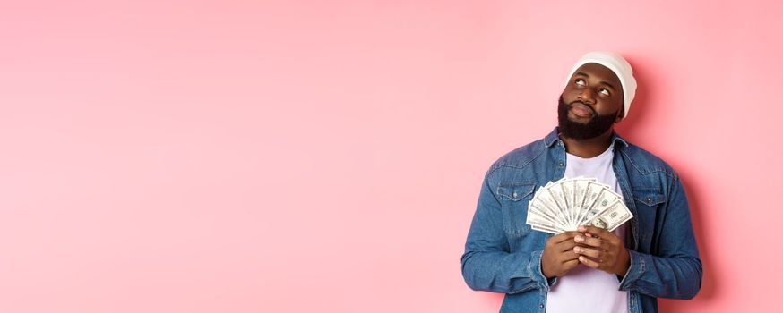 Dreamy african-american man thinking about shopping, holding money dollars and looking upper left corner, standing over pink background.