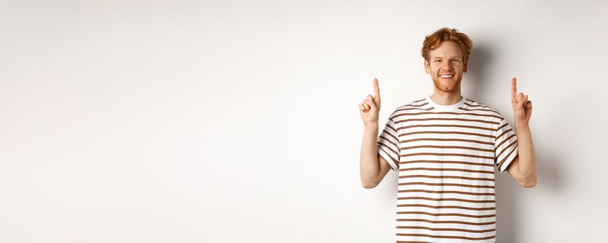 Smiling young male student with red hair showing logo, pointing fingers up and looking happy, standing over white background.