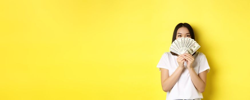 Cute asian woman hiding face behind money, peeking at camera satisfied, earn cash, standing over yellow background.