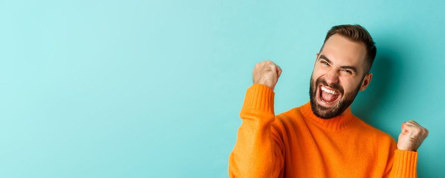 Image of handsome relieved man feeling satisfaction, rejoicing of winning or achievement, making fist pump and saying yes, standing over turquoise background.