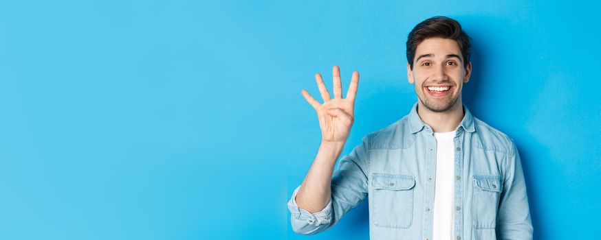Close-up of handsome man smiling, showing fingers number four, standing over blue background.