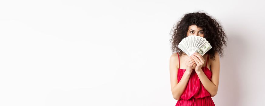 Gorgeous woman with curly hair, hiding face behind dollars and smiling, standing over white background. Copy space