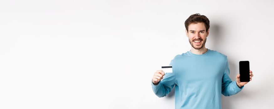 Smiling caucasian man showing plastic credit card with mobile phone screen. Guy recommending online banking app, standing on white background.