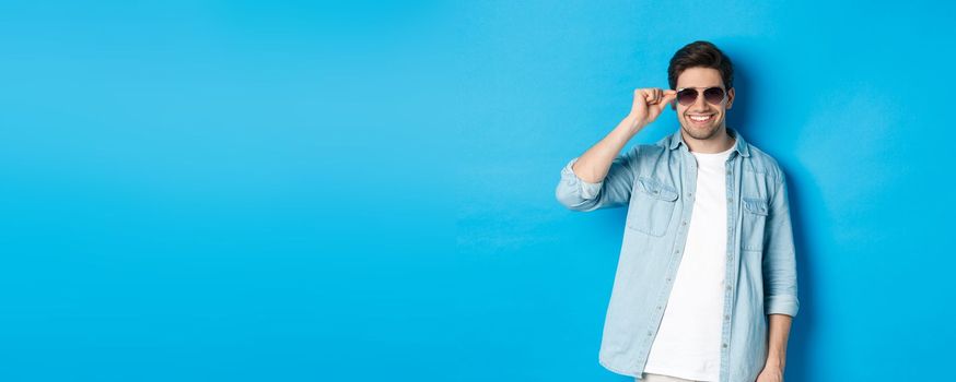 Confident macho man put on sunglasses, looking cool and sassy, standing over blue background.