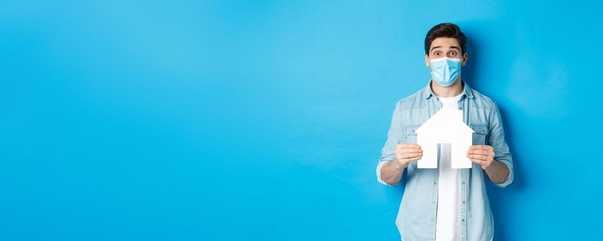 Concept of coronavirus, quarantine and social distancing. Young man searching apartment, showing house paper model, wearing medical mask, renting or buying propery, blue background.