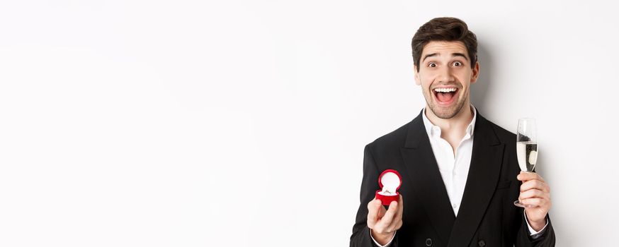 Close-up of handsome man in suit, making a proposal, giving engagement ring and raising glass of champagne, standing against white background.