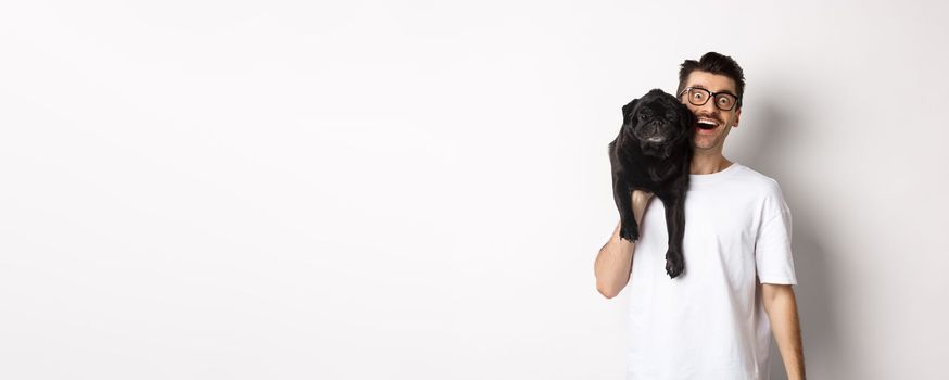 Cheerful hipster guy in glasses and t-shirt, carry cute black pug on shoulder and smiling. Dog owner playing with his pet, standing over white background.