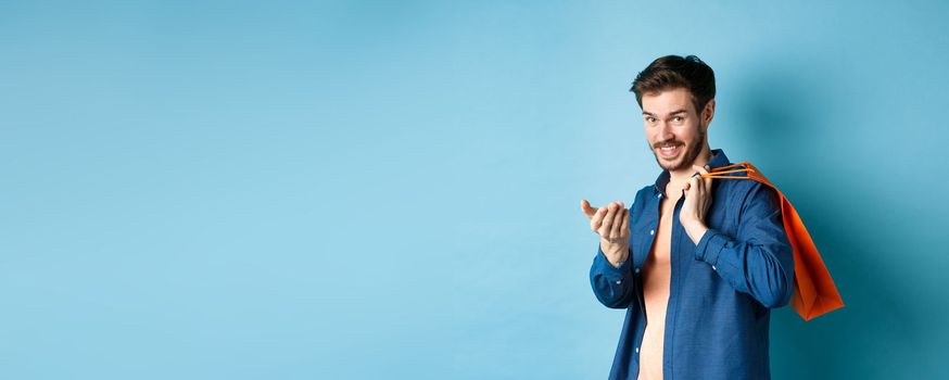 Modern guy holding shopping bag behind shoulder and beckon you, telling to come closer or inviting to store, standing on blue background.