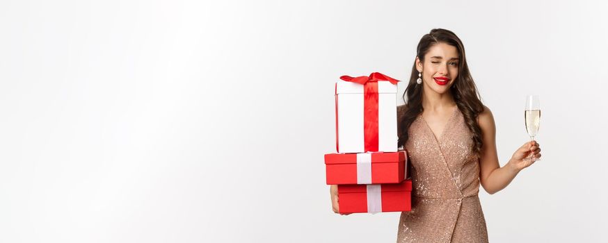 New Year, Christmas and celebration concept. Elegant woman in luxury dress and red lipstick, holding gifts and drinking champagne on party, standing over white background.