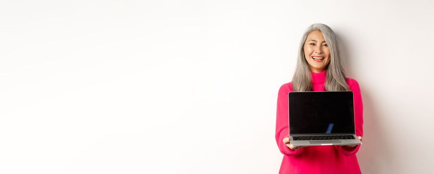 E-commerce concept. Beautiful asian senior businesswoman showing blank laptop screen and smiling, standing in pink sweater over white background.