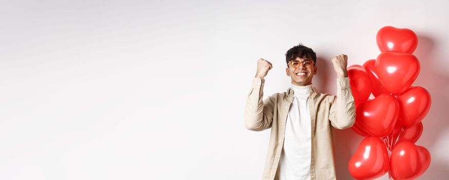 Valentines day. Satisfied young man saying yes, triumphing and celebrating on lovers date, making fist pump and smiling pleased, standing near heart balloons on white background.