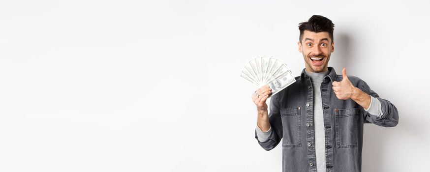Image of excited man holding dollar bills and show thumbs-up with happy face, making money, standing on white background.