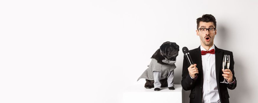 Male entertainer raising glass of champagne, giving microphone to cute black dog, standing over white background.