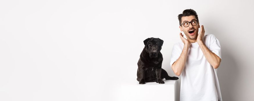 Amazed young man standing with cute black puppy, staring at upper right corner surprised and excited, standing near pug over white background.