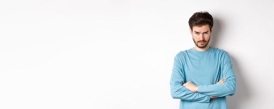 Image of offended and sad young man with beard, look from under forehead and sulking irritated, cross arms on chest defensive, mad at someone, standing on white background.