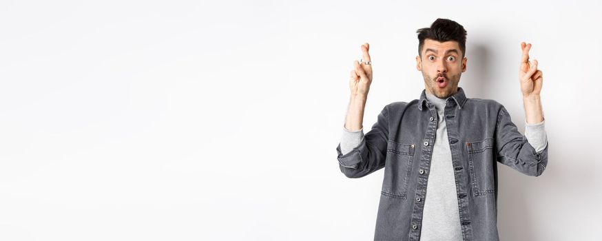 Excited young man cross fingers for good luck and gasping, holding breath as waiting important news, begging or praying, standing on white background.