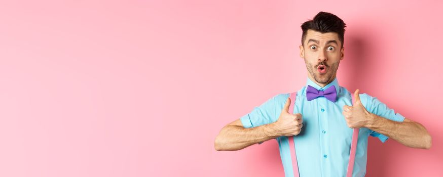 Impressed young guy in suspenders and bow-tie showing thumbs up and saying wow, recommending awesome product, praising quality, standing over pink background.