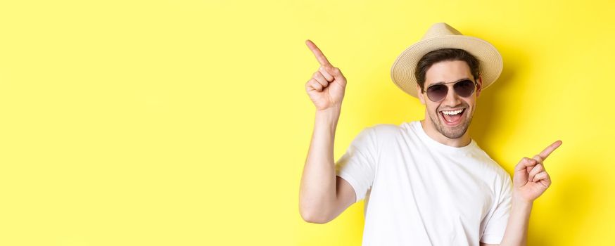 Concept of tourism and vacation. Close-up of man enjoying holidays on trip, dancing and pointing fingers sideways, wearing sunglasses with straw hat.