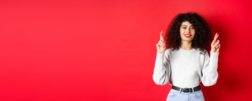 Hopeful young woman with red lips and curly hair, cross fingers for good luck and making wish, praying for dream come true, smiling excited, red background.