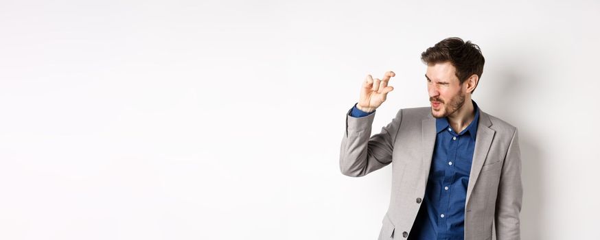 Businessman trying to see something tiny, showing little thing size with fingers, standing in suit on white background.