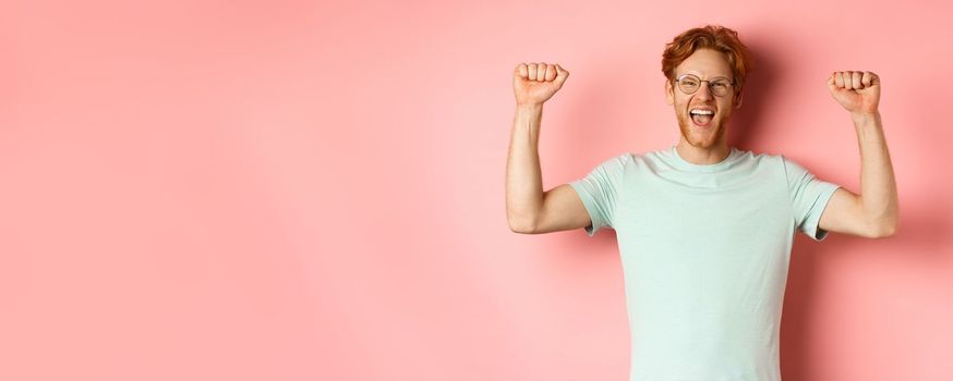 Happy winner with red hair and beard celebrating victory, shouting yes with joy and raising hands up, enjoy success, standing over pink background.
