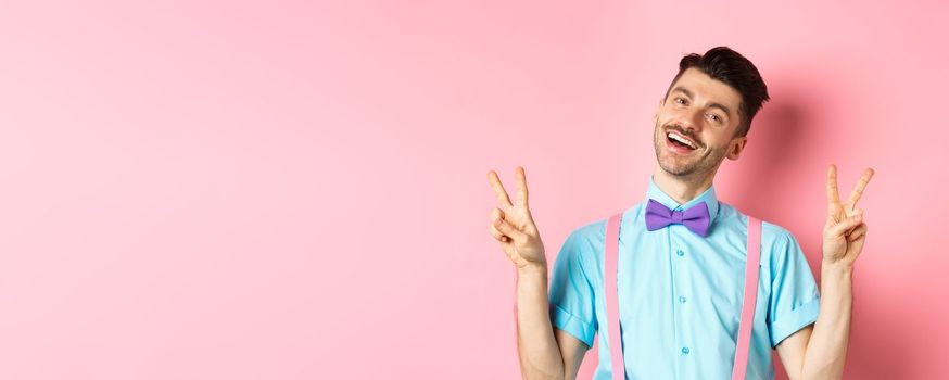 Happy cute guy with moustache and bow-tie, showing peace or victory signs and smiling at camera, posing near pink background.