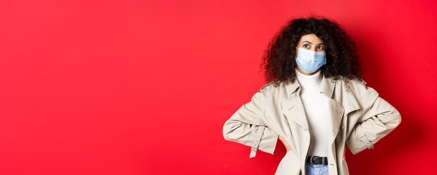 Covid-19, pandemic and quarantine concept. Stylish enthusiastic woman in medical mask and trench coat, smiling and looking at upper left corner, red background.