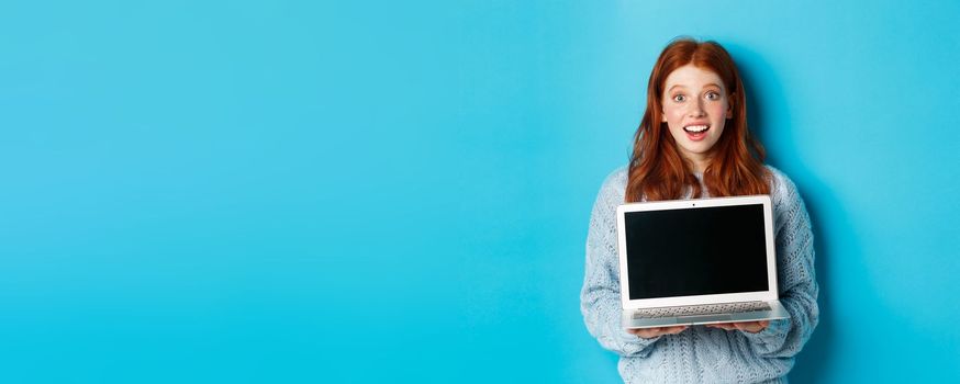 Excited redhead female freelancer showing laptop screen, staring at camera amazed, standing with computer against blue background.