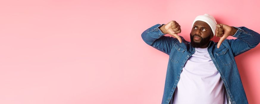 Disappointed Black man staring at upper left corner annoyed, showing thumbs-down, dislike something, standing over pink background.