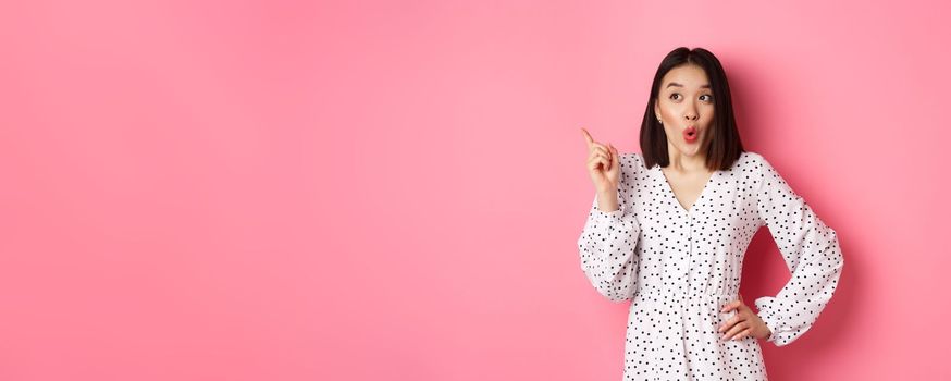 Amazed asian girl in dress pointing finger at upper left corner promo offer, looking interested, shopping and standing over pink background.