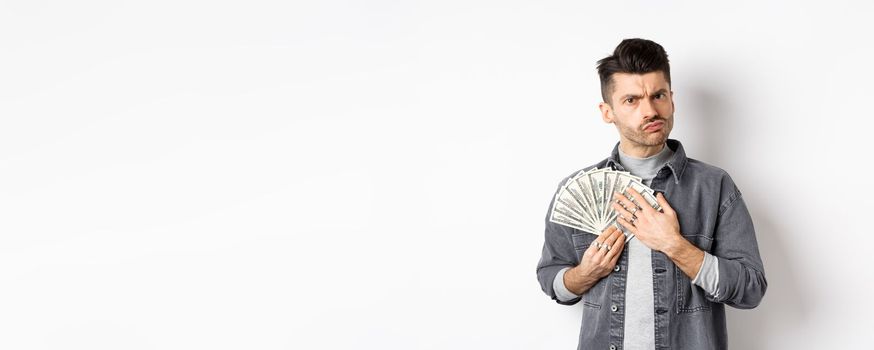 Greedy guy frowning and hugging dollar bills, unwilling to share money, standing on white background.