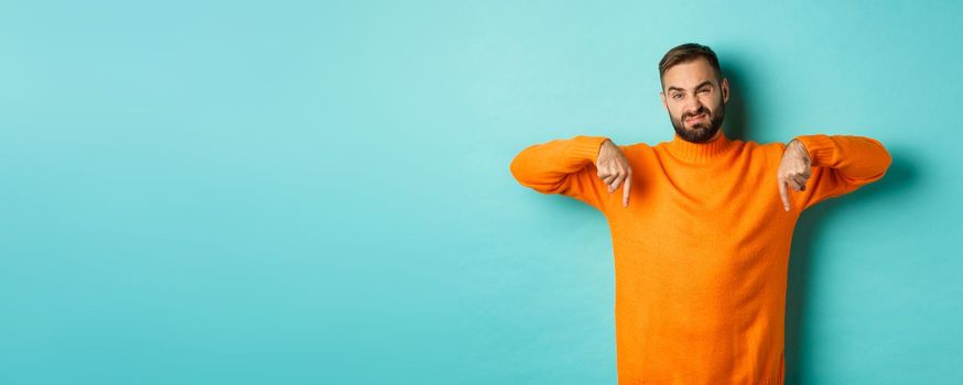 Displeased and skeptical handsome man pointing fingers down, showing bad product, dislike store or advertisement, grimacing disappointed, standing over turquoise background.