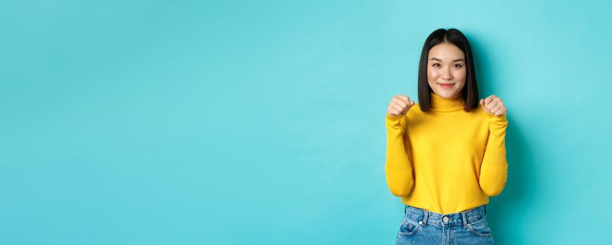 Beauty and fashion concept. Beautiful and stylish asian woman in yellow pullover, holding hands raised near chest as if holding banner or logo, standing over blue background.