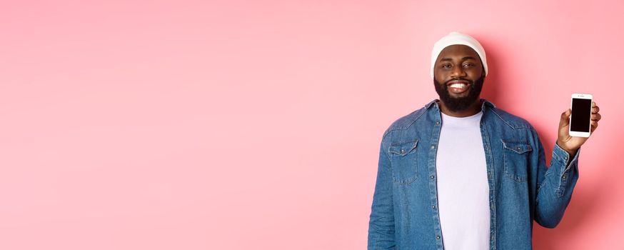 Handsome hipster guy in beanie and denim shirt smiling, showing mobile phone screen with happy face, introduce application, standing over pink background.