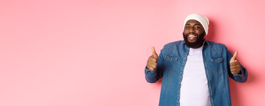 Satisfied handsome black man looking happy, showing thumbs-up and smiling, give approve and praise product, recommending company, pink background.