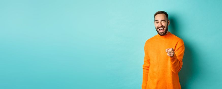 Cheeky attractive man congrats you, praising good job, winking and pointing at camera, standing over light blue background.