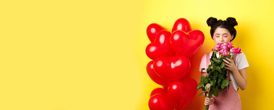 Happy Valentines day. Tender asian girl smelling flower from boyfriend. Girlfriend holding beautiful pink roses, standing near romantic hearts balloons, yellow background.