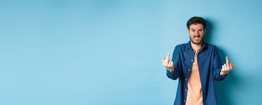 Angry rude guy showing middle finger and saying fuck you, swearing and staring furious at camera, standing on blue background.