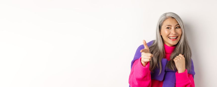 Close-up of happy and cool senior hipster woman, checking out, pointing and looking at person, smiling sideways, standing over white background.