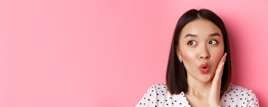 Beauty and lifestyle concept. Close-up of coquettish asian woman looking amazed left, staring at shopping promo offer, standing over pink background.