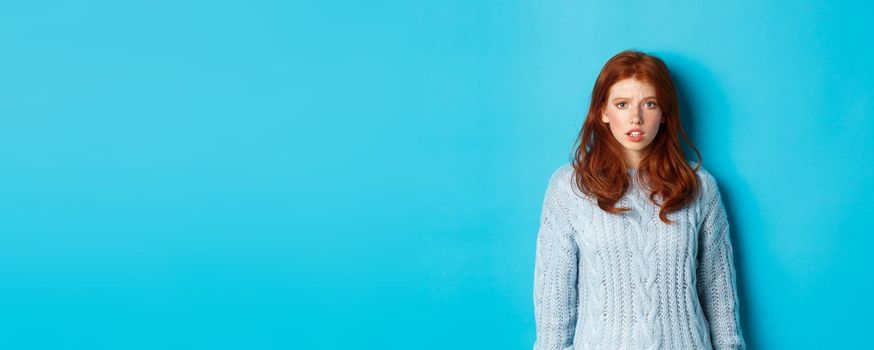 Confused redhead girl in sweater staring at camera, raising eyebrow and feeling puzzled, standing over blue background.