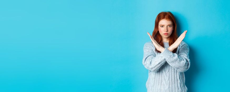 Serious redhead girl looking confident, showing cross gesture to stop and forbid action, standing over blue background.