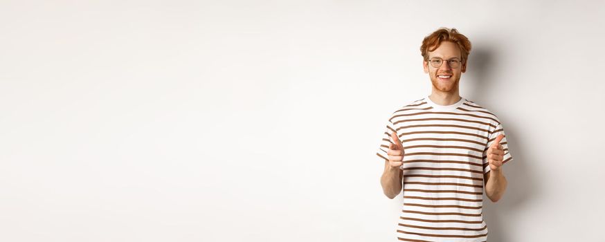 Young man with red hair and glasses pointing fingers at camera and smiling, need you, standing over white background.