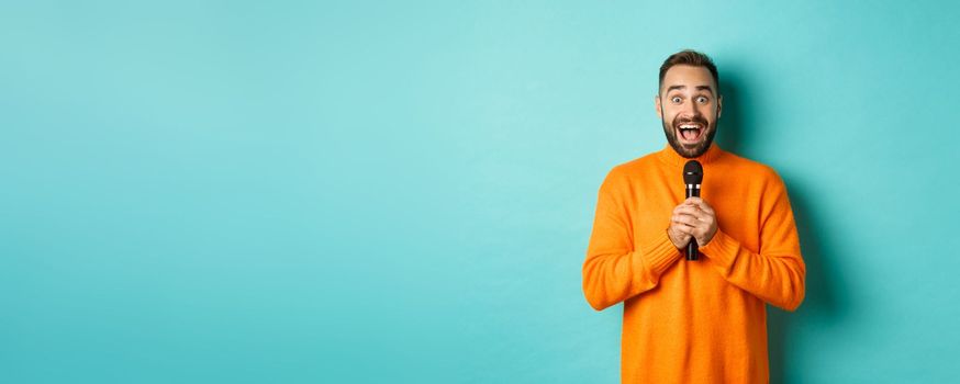 Happy adult man singing karaoke, holding microphone and looking at camera, standing in orange sweater against turquoise background.
