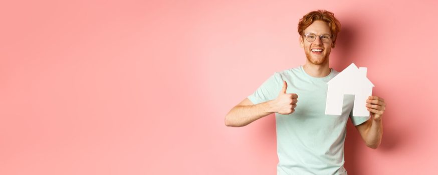 Real estate. Cheerful man in glasses and t-shirt recommending broker agency, showing paper house cutout and thumbs-up, standing over pink background.